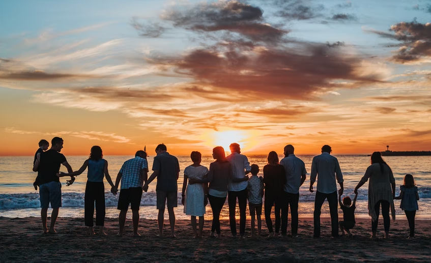 broad demographic of people standing in a line on the beach facing a sunset
